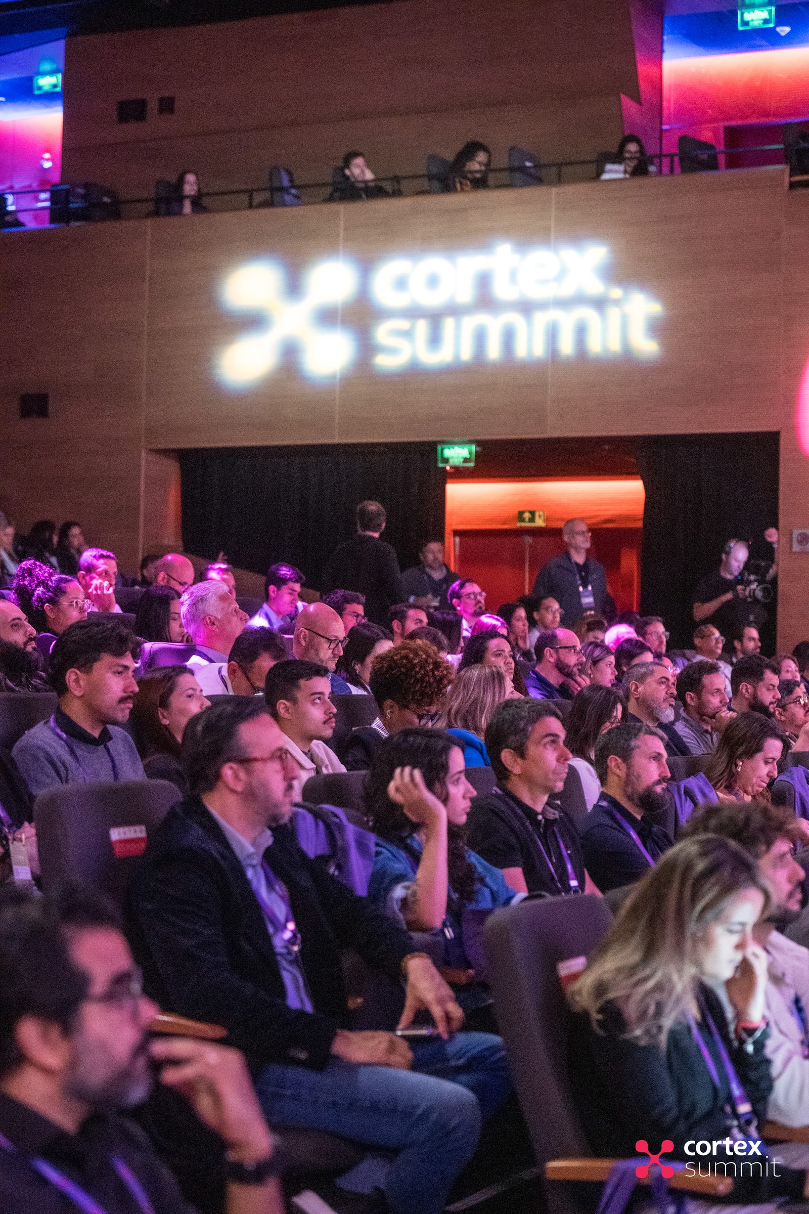 Audience in the theatre facing a stage lit with the Cortex Summit logo