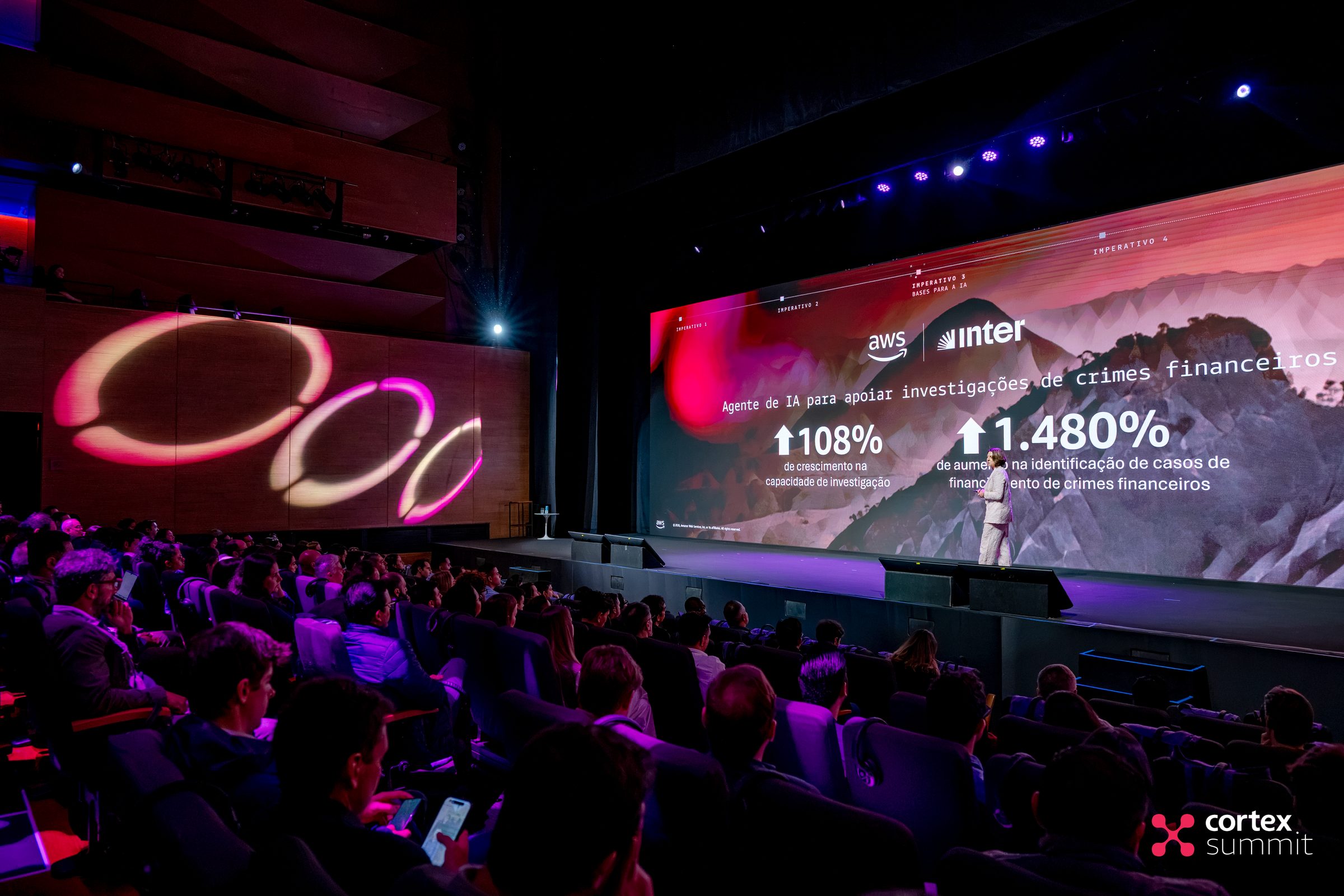 Audience in the dark theatre facing a stage with an outlined circular graphic and keynote data